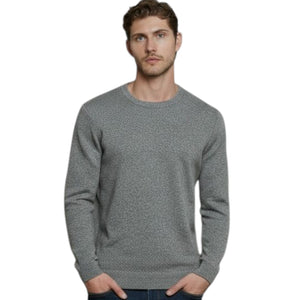 A man stands indoors in front of large windows, wearing the AUTUMN CASHMERE - CASHMERE BASIC CREW NECK WITH RIB BOTTOM IN STONE, paired with dark blue jeans and white sneakers. The cityscape visible outside the windows complements his refined taste in men’s knitwear from AUTUMN CASHMERE.