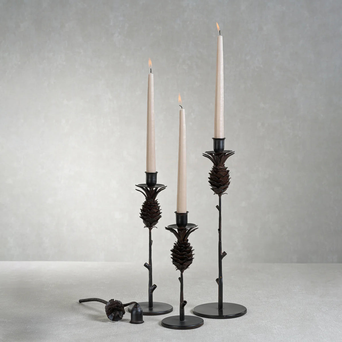 The ZODAX PINE CONE DESIGN WROUGHT IRON CANDLE HOLDER - SHORT features a black pinecone shape on a slender stem with a round base, displayed upright against a white background.