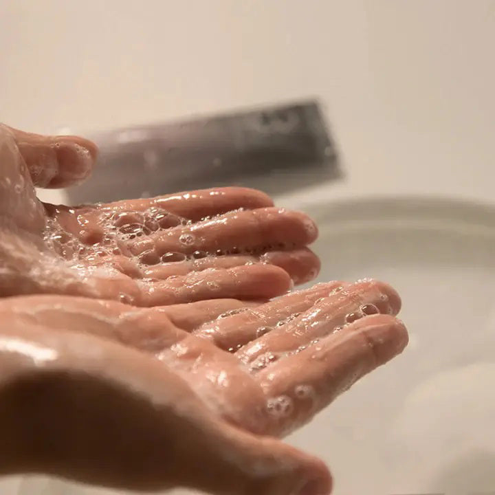 A person squeezes ASIAN BEAUTY WHOLESALE BLACK RICE TRIPLE AHA GENTLE CLEANSING GEL 100ML from a pink and black tube onto their hand over a sink, with a tiled wall in the background. The cleanser uses natural surfactants for gentle care.