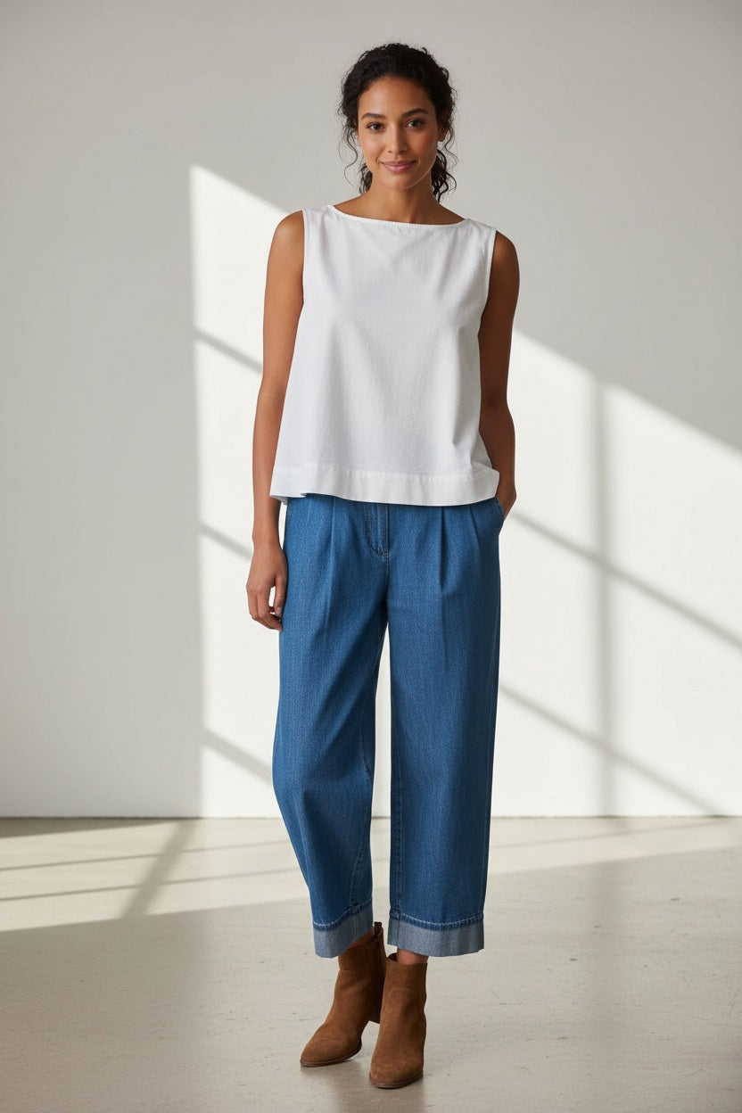 A woman wearing the FRANK AND EILEEN - SHELLEY SHELL TANK IN FAMOUS DENIM WHITE by FRANK &amp; EILEEN and a bright pink skirt stands in front of a white wooden fence, looking down as she touches her hair. A striped bag and towel are on a nearby bench.