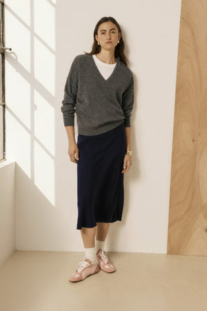 A woman with long brown hair and a neutral expression stands against a two-tone wall, wearing a grey sweater layered over a white shirt, white socks, pink shoes, and the HARTFORD - JIMA Bias Cut Skirt in Night by HARTFORD, effortlessly showcasing chic style with this navy midi skirt.