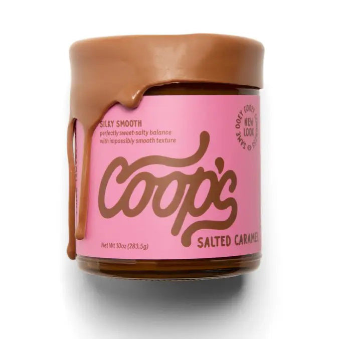 A jar of COOPS - SALTED CARAMEL SAUCE with pink labeling sits open, its thick, silky smooth sauce dripping down the side. The label reads "COOPS," boasting an all-natural, gluten-free treat with a perfectly sweet-salty balance and impossibly smooth texture.