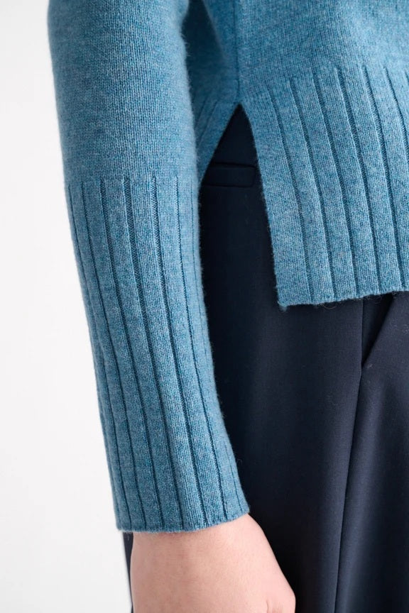 A woman with blonde hair models the KINROSS CASHMERE Hi Low Rib Trim Funnel in Loch—a light blue, long-sleeve cashmere pullover with ribbed trim—paired with dark pants. She stands against a plain white background, turned slightly to the side.