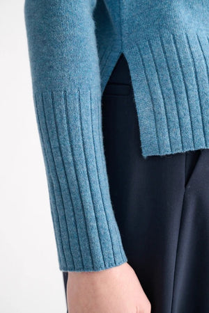 A woman with blonde hair models the KINROSS CASHMERE Hi Low Rib Trim Funnel in Loch—a light blue, long-sleeve cashmere pullover with ribbed trim—paired with dark pants. She stands against a plain white background, turned slightly to the side.