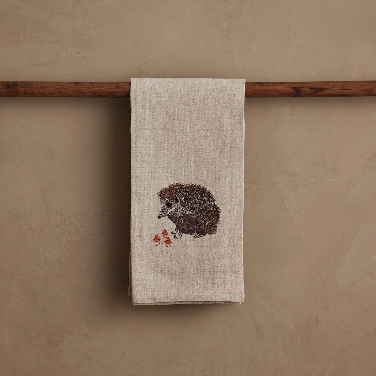 A HEDGEHOG WITH MUSHROOMS LINEN TEA TOWEL by CORAL & TUSK hangs from the handle of a white stove in a kitchen.