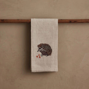 A HEDGEHOG WITH MUSHROOMS LINEN TEA TOWEL by CORAL & TUSK hangs from the handle of a white stove in a kitchen.