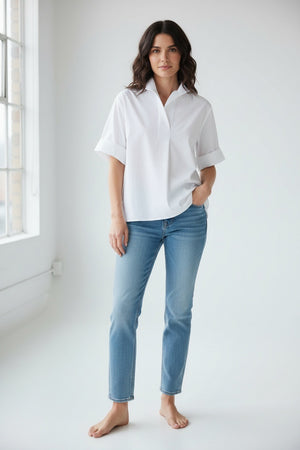 A woman with short light brown hair is seen standing against a plain light background, wearing the HINSON WU - CARA Elbow Sleeve Shirt in Stretch Polished Cotton White by HINSON WU. The oversized, loose-fit shirt features a polished cotton fabric with a collared neckline and elbow-length sleeves, and is paired with light blue jeans, large hoop earrings, and a gold bracelet. The machine-washable shirt offers effortless care.