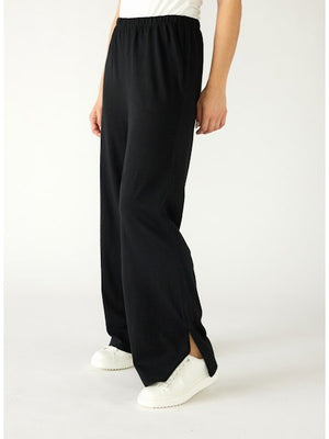 A woman stands in a minimal white room wearing the MERSEA TRAVELER TEE PANT, light gray, made from Turkish cotton slub—styled with white sneakers featuring green accents—and smiles softly at the camera.