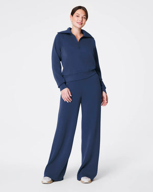 A woman stands against a plain white background, modeling the SPANX - AIRESSENTIALS WIDE LEG PANT in dark blue, paired with a long-sleeved, half-zip pullover made from lightweight fabric. She has her right hand touching the zipper near her chin and is wearing white sneakers. The relaxed fit adds to the ensemble's casual appeal.