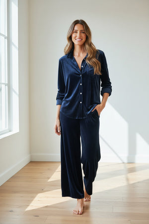 A woman with long, wavy hair smiles while wearing the FRANK & EILEEN – EILEEN Relaxed Button Up in Navy Velvet. This shirt’s relaxed fit creates a bust-flattering silhouette, embodying an effortlessly casual California vibe as she stands against a plain light background with her hands in her pockets.