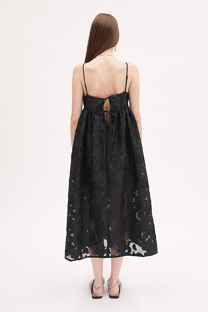 A woman wears the MEIMEIJ Lace Empire Waist Dress, a sleeveless black floral lace midi with spaghetti straps and a sheer overlay. The flowing fit makes it perfect for a garden party. She stands against a plain white background.