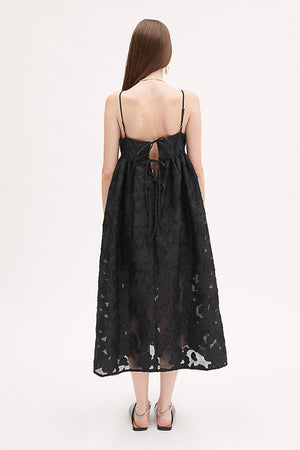 A woman wears the MEIMEIJ Lace Empire Waist Dress, a sleeveless black floral lace midi with spaghetti straps and a sheer overlay. The flowing fit makes it perfect for a garden party. She stands against a plain white background.