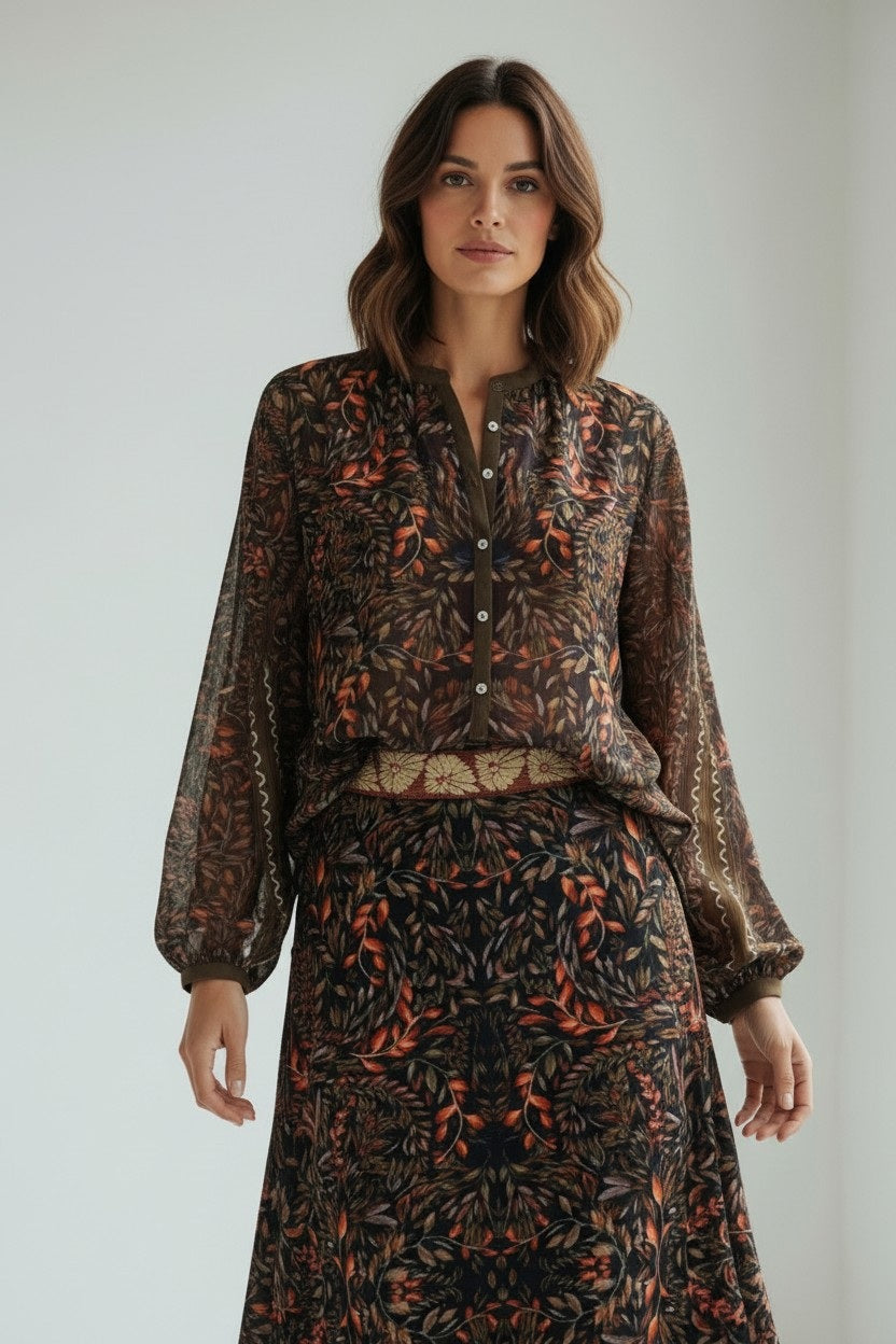 A woman with shoulder-length brown hair and a neutral expression stands indoors wearing the FRANCO FERRARI GERRY Silk Top, a sheer, long-sleeved piece featuring a vibrant botanical print, paired with a matching skirt.