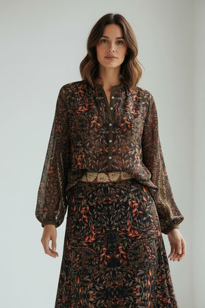 A woman with shoulder-length brown hair and a neutral expression stands indoors wearing the FRANCO FERRARI GERRY Silk Top, a sheer, long-sleeved piece featuring a vibrant botanical print, paired with a matching skirt.