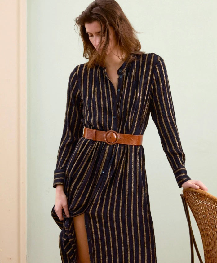 A woman in the MOMONI Aubert Dress in Lurex Gauze stands by a woven chair, gazing down as her hair partially covers her face.