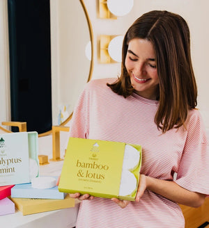 A green box labeled “MUSEE - BAMBOO & LOTUS SHOWER STEAMERS” is displayed, featuring four white round steamers embossed with a leaf design in front. The brand name “MUSEE” appears above the product name, highlighting these natural shower steamers crafted to deliver a spa-like experience.