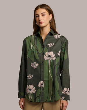 A woman with long brown hair wears the HINSON WU Halsey Long Sleeve Shirt in Poppy Print and tan pants, standing with her hands in her pockets against a neutral background.