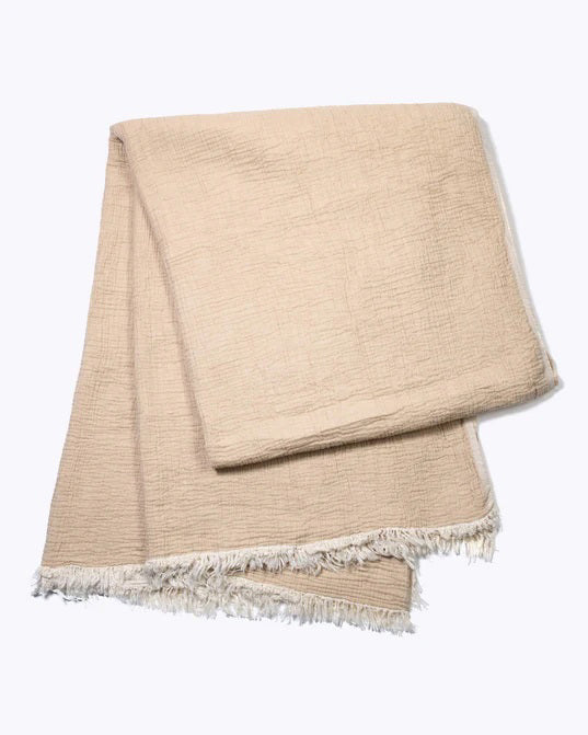 The MERSEA CLOUD GAUZE BLANKET, shown folded on a light background, is a soft beige cotton throw with a textured surface and fringed edges.
