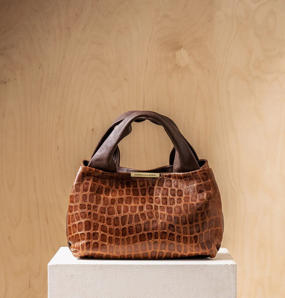 The DANIELLA LEHAVI EVELINE Satchel in Brown Mosaic, featuring double handles, a croco-inspired texture, and an adjustable shoulder strap, hangs on a wall hook against a light wooden background.