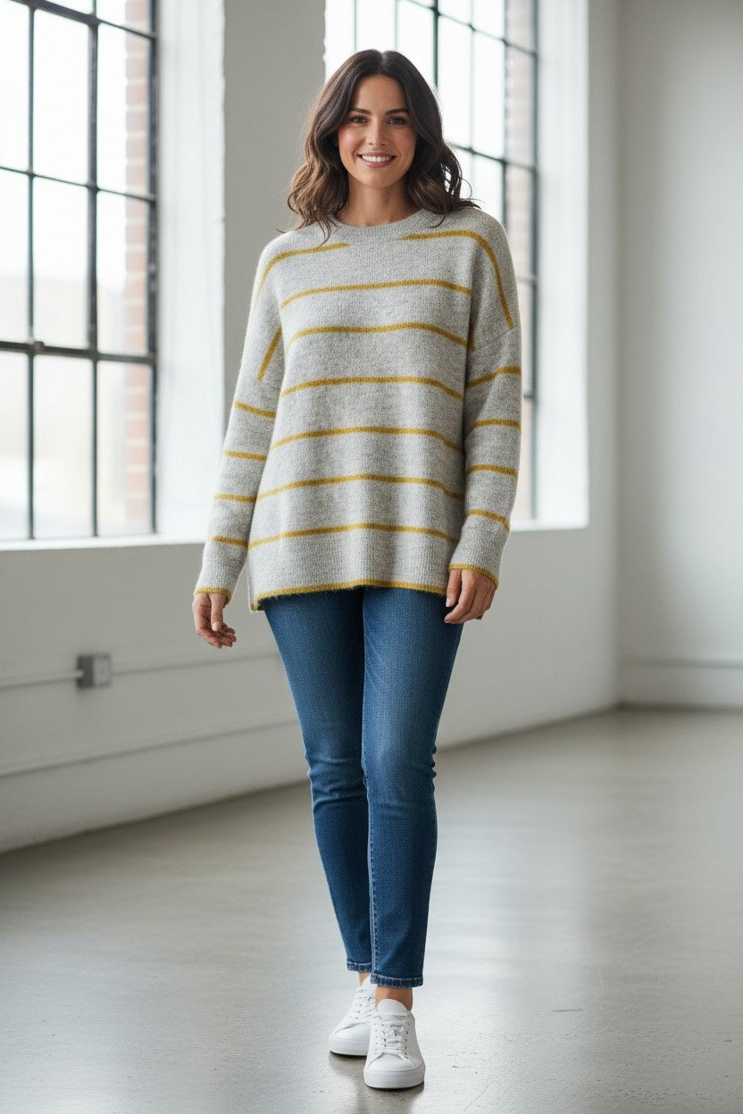 A woman stands indoors near large windows, smiling. She wears the & ISLA Lofty Oversized Stripe Sweater in Frost/Moss—an eye-catching knit featuring yellow stripes—paired with blue jeans and white sneakers. The room has light-colored walls and a polished concrete floor.