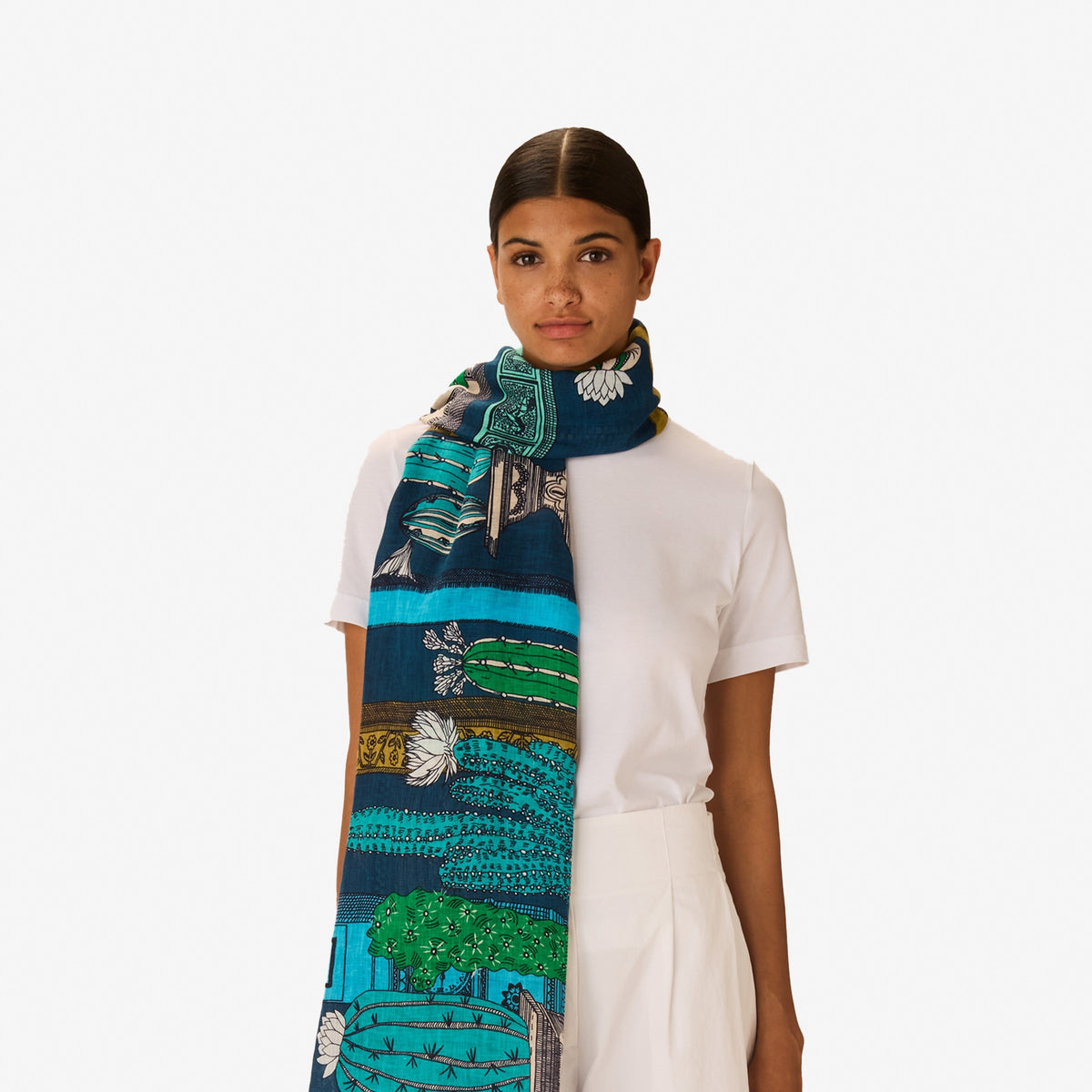 A woman with dark hair tied back wears a white outfit, accessorized with the INOUI EDITIONS - TEQUILA SCARF IN CURACAO by INOUI, featuring green, yellow, and blue patterns, set against a plain white background.