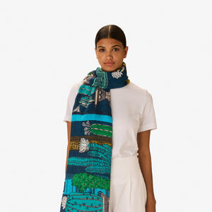 A woman with dark hair tied back wears a white outfit, accessorized with the INOUI EDITIONS - TEQUILA SCARF IN CURACAO by INOUI, featuring green, yellow, and blue patterns, set against a plain white background.