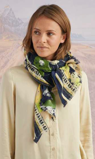 A woman outdoors with a blurred mountain backdrop wears a cream button-up and the STORIATIPIC DORIS Wool Silk Scarf by STORIATIPIC, featuring green, navy, and yellow patterns in a luxe wool-silk blend.