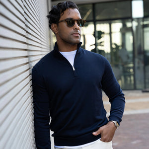 A man in the COLLARS AND CO Maranello Cotton & Silk Quarter Zip in Navy, sunglasses on, leans against a striped wall outside with one hand in his pocket and a modern glass building behind him, capturing elevated casual style.