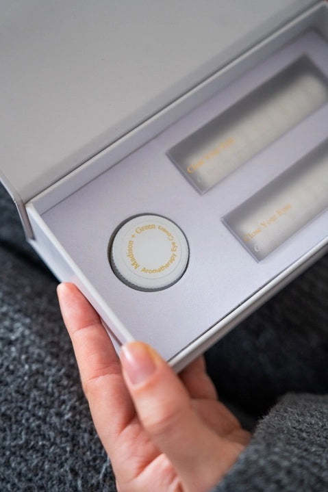 A person in a gray sweater holds an open white box containing MADISON GREEN’s “REST YOUR EYES ON THE COUCH,” lifting the small jar with a white and gold lid from the box—perhaps an essential oil blend designed for calming eye masks or aromatherapy eye covers.