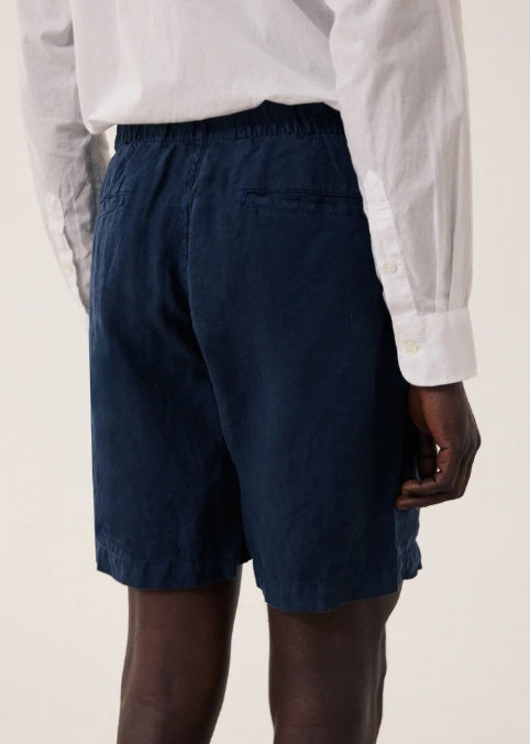 A person wearing a white long-sleeve shirt, HARTFORD - LINEN JOGGY SHORT IN NAVY by HARTFORD, and beige sneakers stands on a wooden floor against a plain light background.