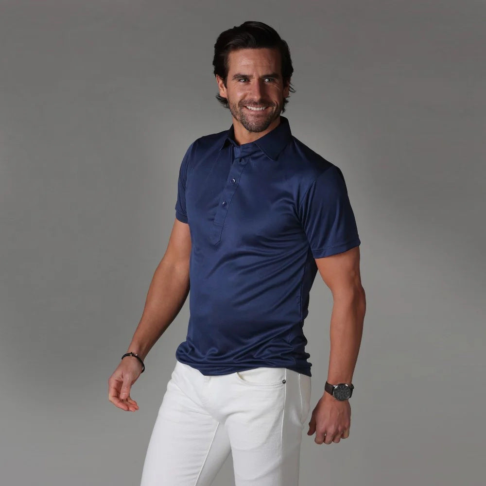 A dark-haired, bearded man smiles while wearing the COLLARS AND CO - SEMI SPREAD COLLAR POLO IN NAVY and white pants, standing against a plain gray background.
