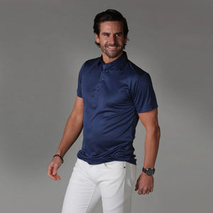 A dark-haired, bearded man smiles while wearing the COLLARS AND CO - SEMI SPREAD COLLAR POLO IN NAVY and white pants, standing against a plain gray background.
