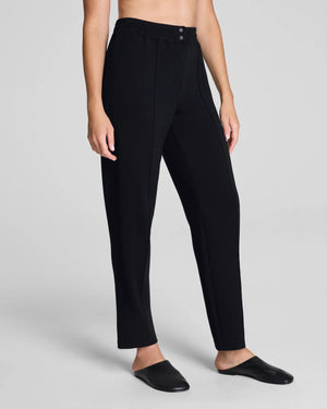 A person models the SPANX - AIRessentials Brushed Slim Straight Pant with an elastic waistband and buttons, paired with black slip-on shoes against a plain light background.