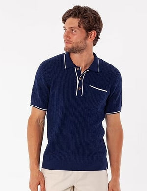 A close-up of a person wearing the CRIQUET Bungalow Sweater Polo in Navy, featuring a textured vintage style, collar, three brown buttons, white trim, and a small breast pocket outlined in white.