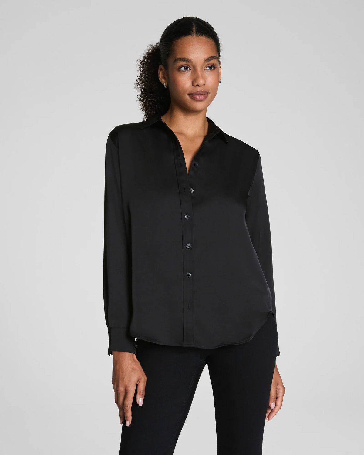 A woman with curly hair pulled back wears the SPANX Silky Smooth Button Down Shirt in black, paired with black pants. She stands facing away, looking over her shoulder against a plain light gray background.