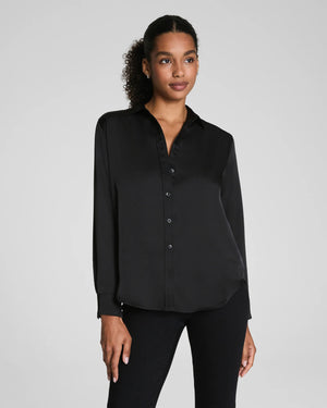 A woman with curly hair pulled back wears the SPANX Silky Smooth Button Down Shirt in black, paired with black pants. She stands facing away, looking over her shoulder against a plain light gray background.