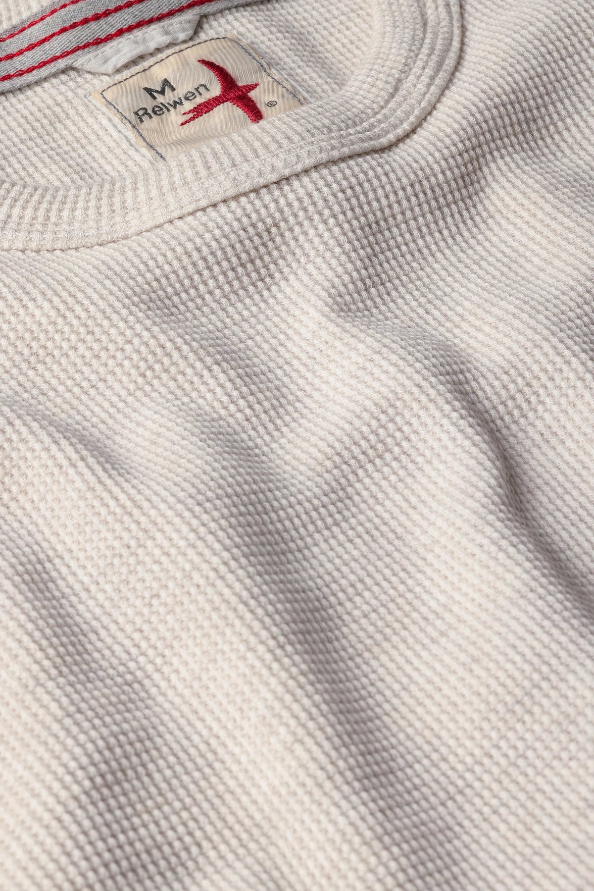 Close-up of the RELWEN Bi-Waffle Crew in Oatmeal, a textured off-white thermal shirt made from 100% cotton and featuring a visible collar along with a fabric tag labeled “RELWEN M” stitched with red thread.