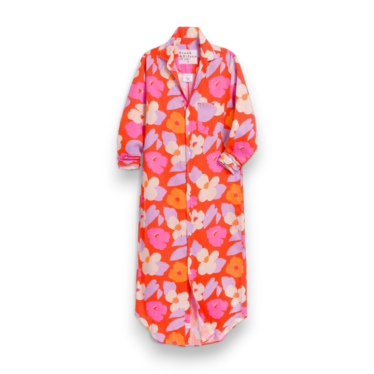 A person with curly hair stands outdoors, wearing the FRANK &amp; EILEEN Rory Maxi Shirtdress in Classic Italian Linen—Orange and Pink Floral—embodying California chic amid lush greenery and white outdoor furniture, holding a woven straw bag.