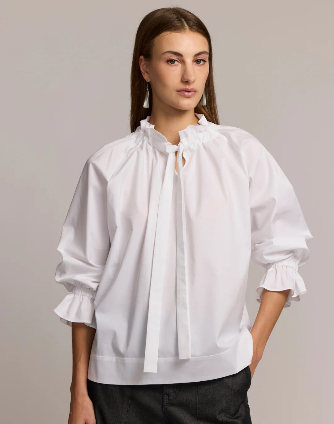 A woman with long brown hair models the HINSON WU ALINA LONG SLEEVE SHIRT, a bright blue blouse with a bow neck and ruffled sleeves, paired with dark pants. She stands against a light background, looking at the camera.