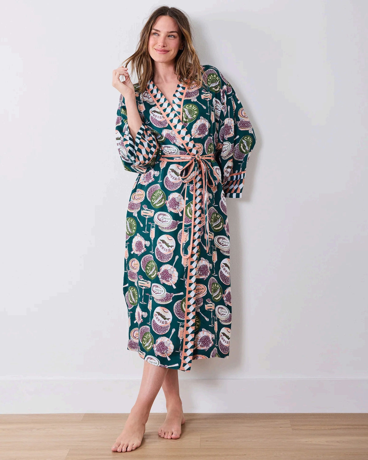 A woman stands barefoot against a white wall, smiling and holding a white cup in one hand as she poses slightly angled to the side. She is wearing the PRINTFRESH Champagne & Caviar Eco Satin Robe in Peacock Feather—a long, colorful eco-friendly robe featuring a vibrant botanical print with peacock feathers and checkered trim.