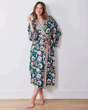 A woman stands barefoot against a white wall, smiling and holding a white cup in one hand as she poses slightly angled to the side. She is wearing the PRINTFRESH Champagne & Caviar Eco Satin Robe in Peacock Feather—a long, colorful eco-friendly robe featuring a vibrant botanical print with peacock feathers and checkered trim.