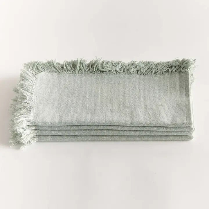 A stack of NAPA HOME AND GARDEN - AURA DINNER NAPKIN IN LIGHT GREEN, featuring fringed edges, is folded and placed on a white surface.