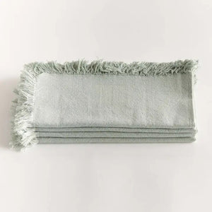 A stack of NAPA HOME AND GARDEN - AURA DINNER NAPKIN IN LIGHT GREEN, featuring fringed edges, is folded and placed on a white surface.