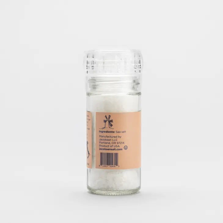 A clear plastic grinder of JACOBSEN SALT CO. GRINDER COARSE ITALIAN SEA SALT sits on a light surface with a plain gray background. The grinder features an orange label with black text.