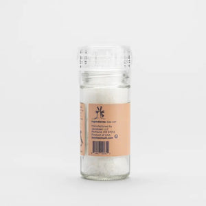 A clear plastic grinder of JACOBSEN SALT CO. GRINDER COARSE ITALIAN SEA SALT sits on a light surface with a plain gray background. The grinder features an orange label with black text.
