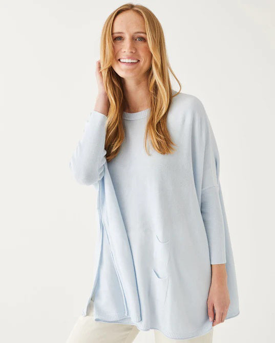 A smiling woman with curly hair wears the MERSEA - CATALINA CREWNECK SWEATER in navy blue and jeans, standing against a plain light background with her hands near her head.