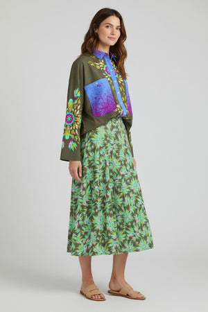 A woman with long brown hair, smiling slightly, wears a loose-fitting, long-sleeve top with colorful patterns, the FRANCO FERRARI Floral Print Skirt from Italy in green, and beige sandals.