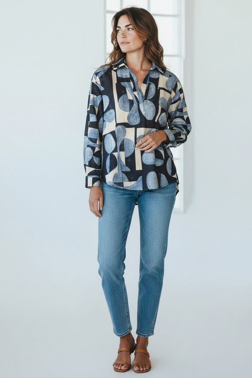 A woman with wavy brown hair stands in a bright room, exuding effortless French elegance as she looks to the side. She wears the HARTFORD - CHORUS POPOVER SHIRT IN BLUE by HARTFORD, styled with light blue jeans and brown sandals. The shirt features a patterned long sleeve design and a chic shirttail hem.