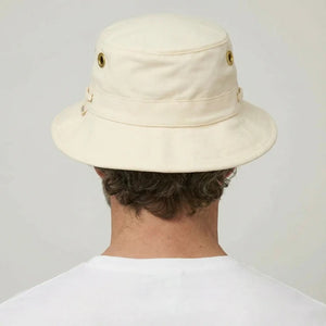 A bearded man in a white shirt wears the TILLEY - ICONIC T1 BUCKET HAT IN NATURAL with UPF 50+ protection and metal eyelets, looking directly at the camera against a plain background.