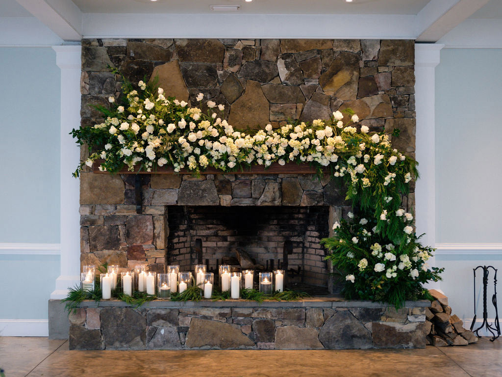 A rustic stone fireplace decorated with lush white flowers and greenery, with numerous lit candles arranged along the hearth. Firewood is stacked on the right side of the fireplace.